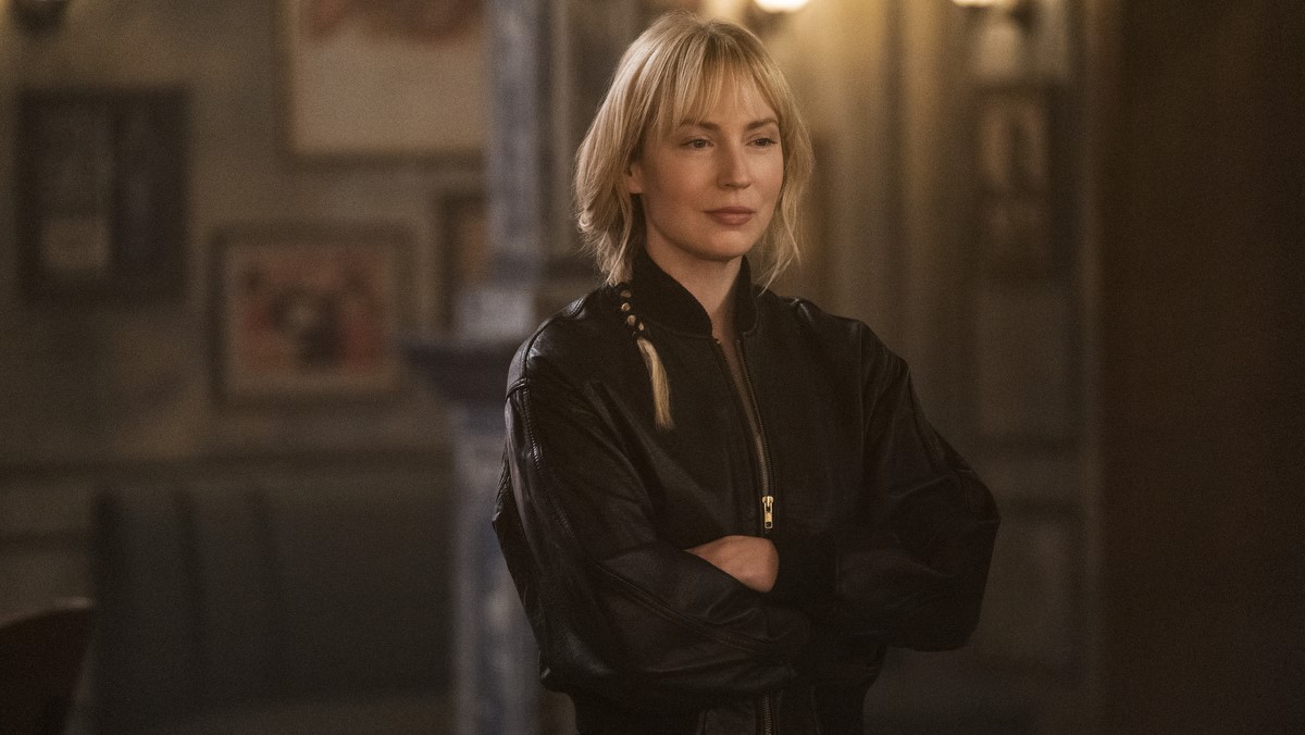 Parker stands in a warehouse with her arms crossed, wearing a black windbreaker jacket.