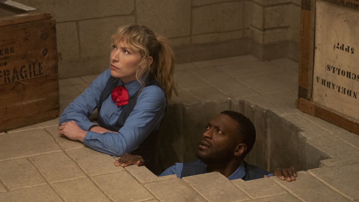 Parker and Hardison peek out of a hole in the floor of a vault dressed in matching uniforms with a black vest over a blue shirt and a red neckerchief. Leverage: Redemption "The Weekend in Paris Job" Episode 301