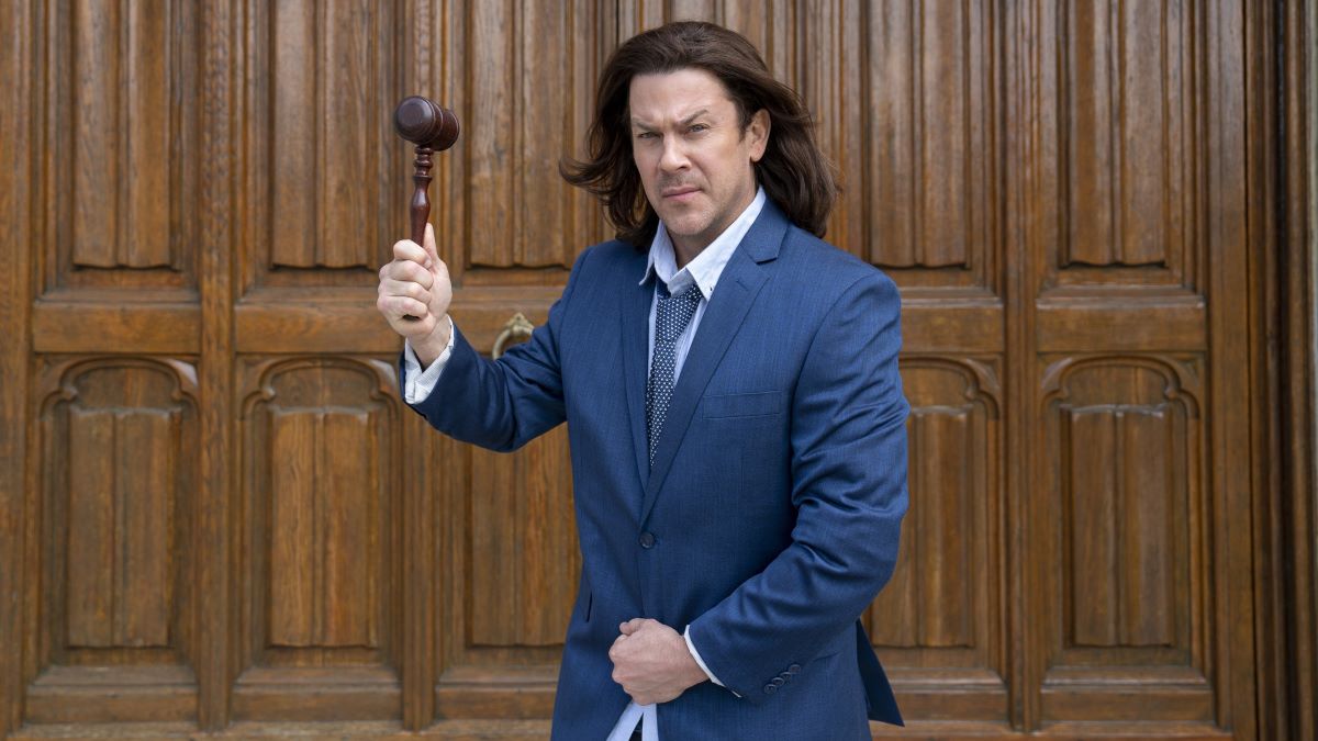 Eliot stands in front of a wood panelled wall with a scowl on his face, a gavel raised in his right hand, holding his blue suit jacket closed with his left. Leverage: Redemption Season 3
