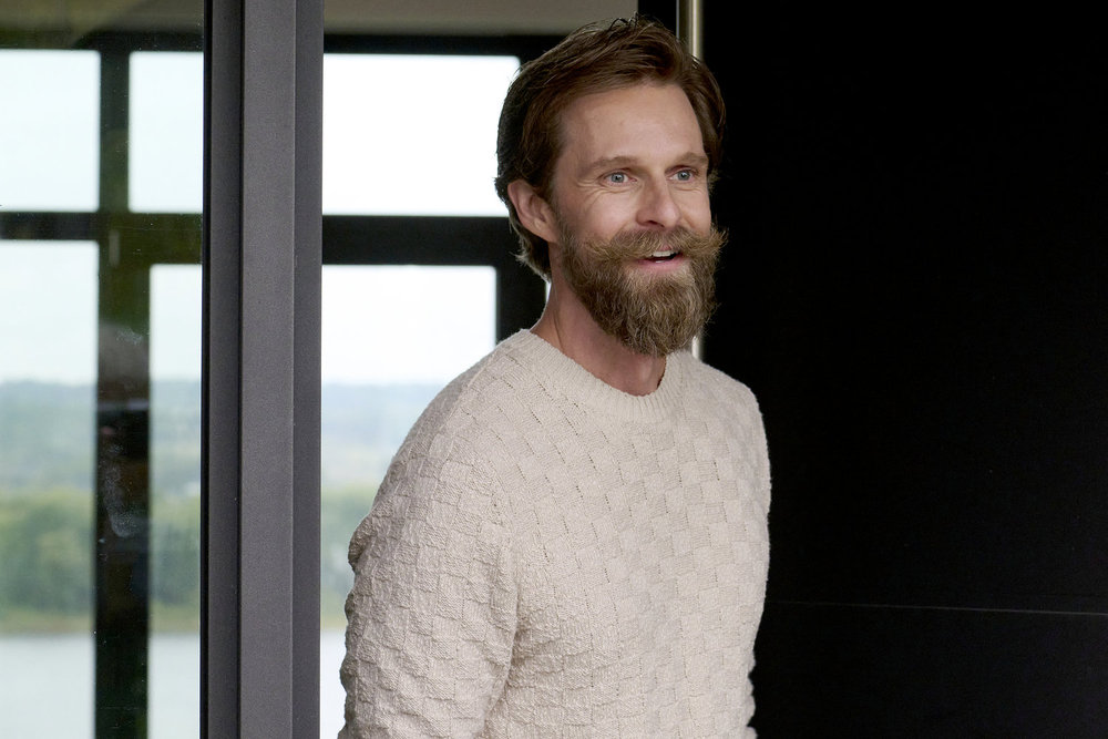 Tyler, a white man, grins while standing on the doorstep of his lavish home. He wears an off-white sweater with a blonde beard and slicked back blonde hair.