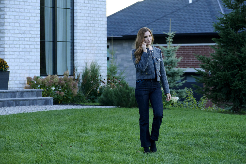 Susan, a white woman with long red hair, walks through a green yard before a lavish house while talking on the phone.
