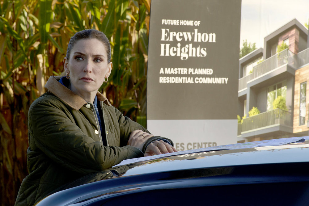 Susan, a white woman with auburn hair, leans on a car while standing outside on a farm during the day.