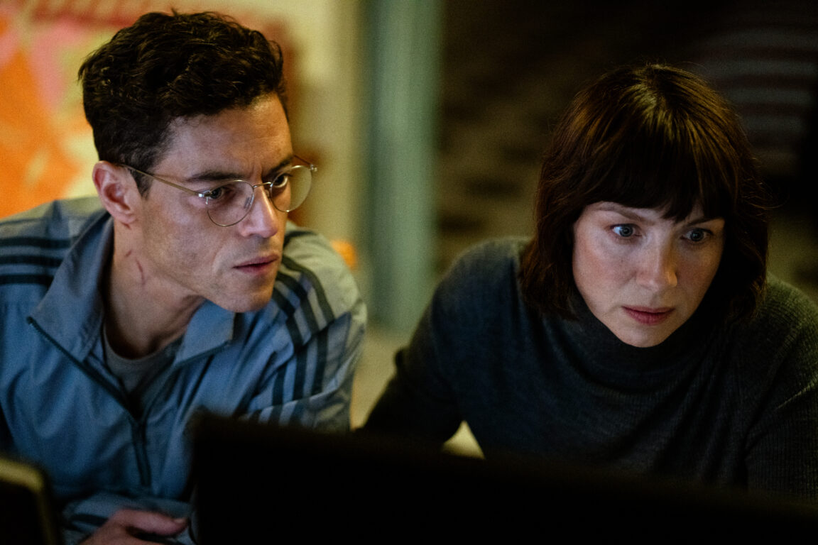 Rami Malek looks over Caitriona Balfe's shoulder as she works on a computer.