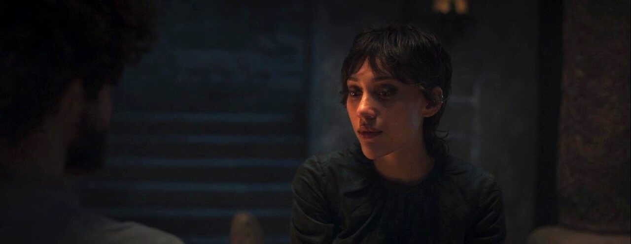 A closeup of Isabella Bucceri as Faile in The Wheel of Time Season 3. She sits in a dimly lit inn before a blazing fire at night.