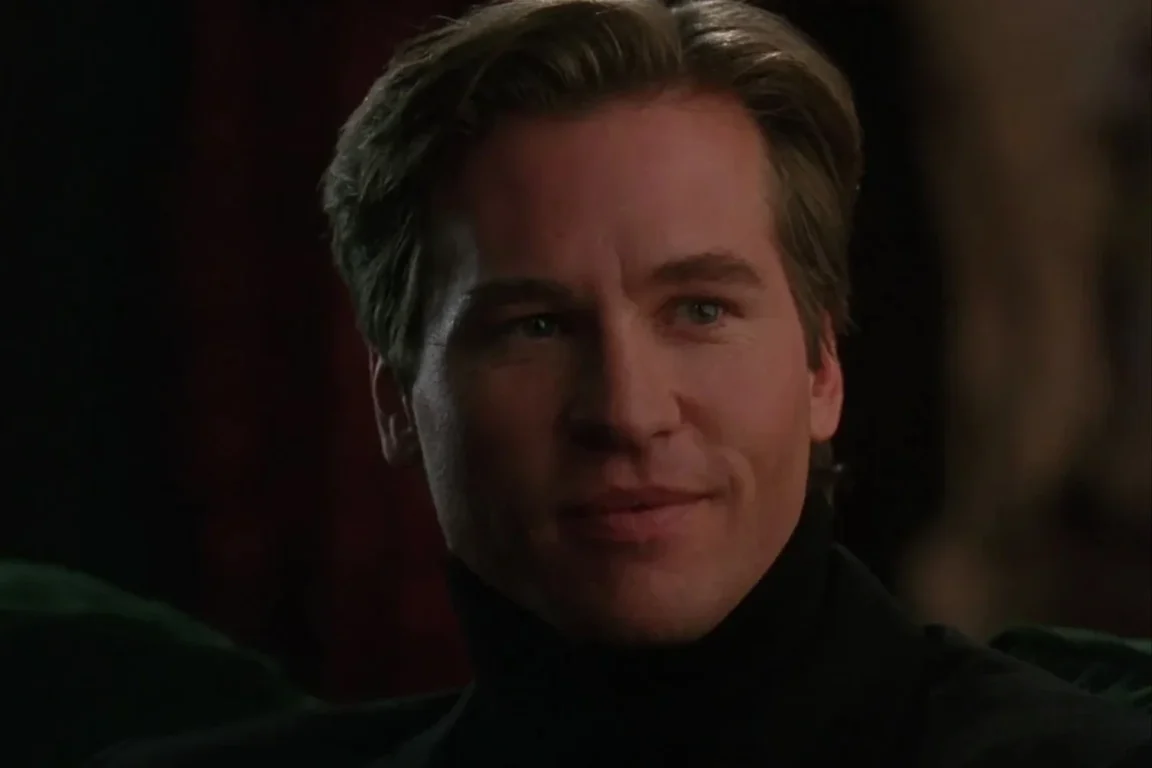 A closeup of actor Val Kilmer. He has short blonde hair and wears a black turtleneck. He smiles softly at someone out of frame.