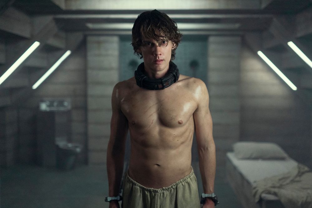 Tyler Galpin stands in a cell with a collar around his neck, shirtless, and wearing tan pants.