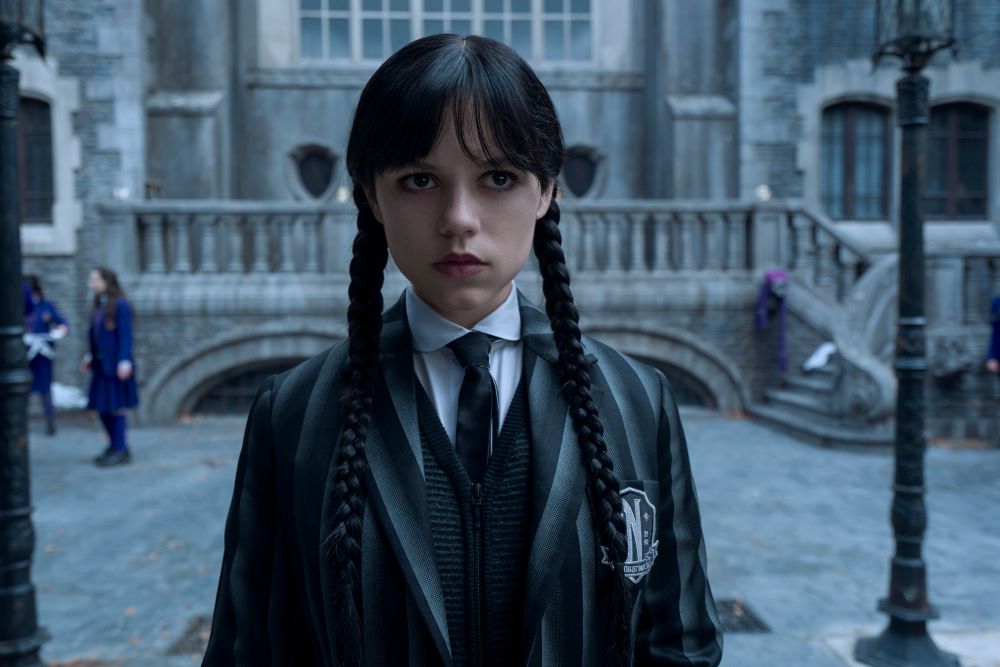 A closeup of Wednesday in her Nevermore Academy uniform as she stands outside the school grounds. She looks focused.