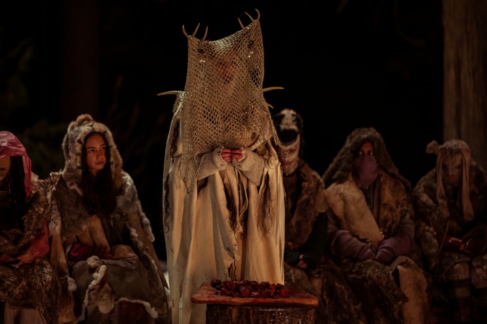 The Antler Queen stands between the teens as they sit around a roaring fire in the snowy woods at night on Yellowjackets Season 3 Episode 10, "Full Circle."