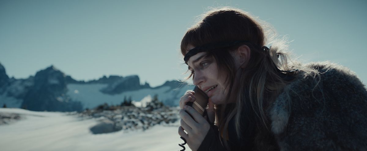 Natalie, a white teen girl, cries while talking on a sat phone as she sits at the top of a snowy mountain range in the Canadian wilderness during the day.