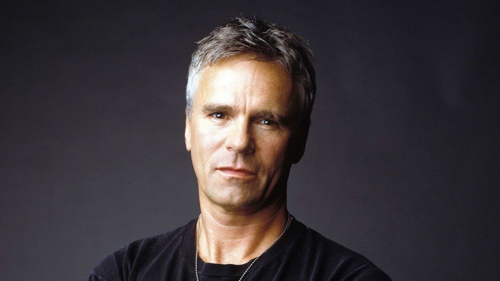 Richard Dean Anderson wears a black t-shirt and stands in front of a gray backdrop with his arms crossed and a dogtag chain around his neck