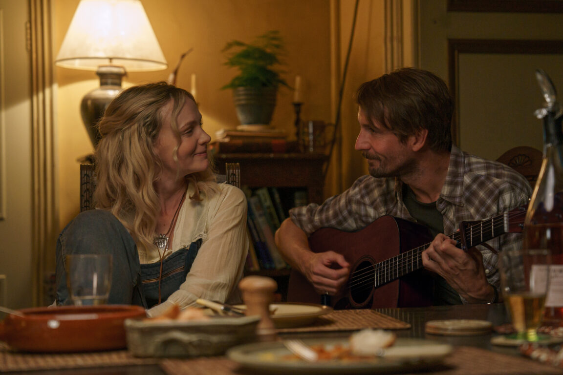 Carey Mulligan and Tom Basden sit at a table and smile at each other. Basden holds a guitar in the movie The Ballad of Wallis Island.