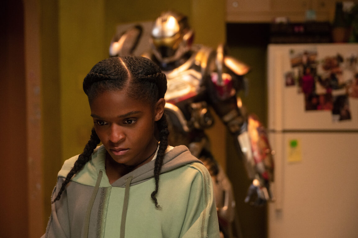 Riri Williams wears a gray hoodie while standing in a dimly lit apartment kitchen. Her Ironheart suit is behind her.