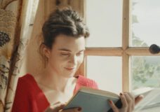 A white woman with brown hair pulled back into a bun sits in a windowsill while reading a book in a still from the movie Jane Austen Wrecked My Life.