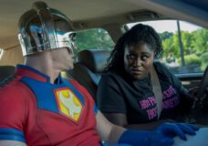 Christopher Smith wears his Peacemaker costume while sitting in the front passenger seat of the vehicle Leota Adebayo drives. Leota looks at him encouragingly.