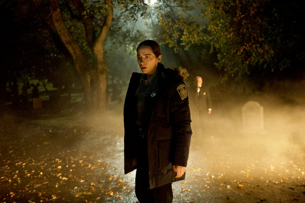Dana Cypress wears a police uniform while standing outside at night. Fog surrounds her in the TV series Revival.
