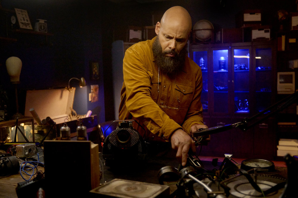 August stands before his workstation in his dimly lit office. He tinkers with a device on SurrealEstate Season 3 Episode 7, "A Slice of Afterlife."