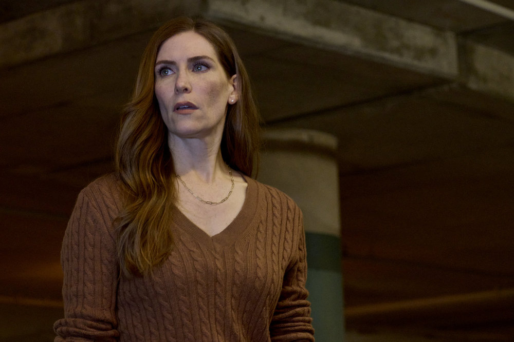 Susan, a white woman with long auburn hair, stands in a parking garage at night. She looks concerned.