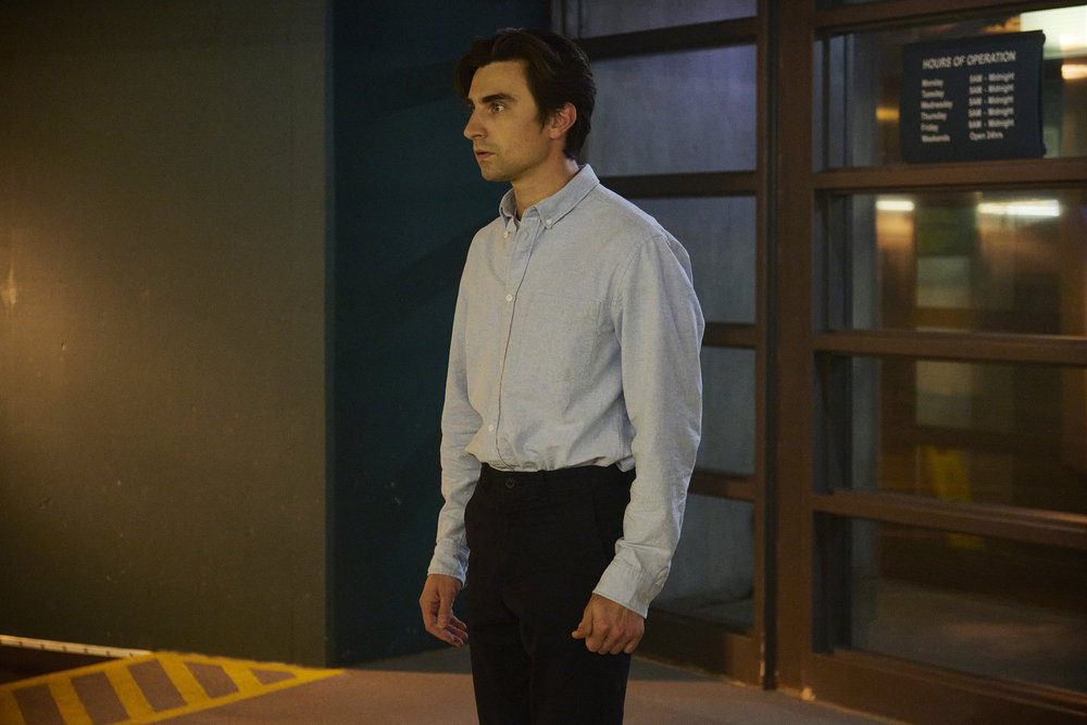 Tag, a young white man with brown hair, stands in a parking garage at night while looking focused. He wears a light blue button-down shirt and black slacks.