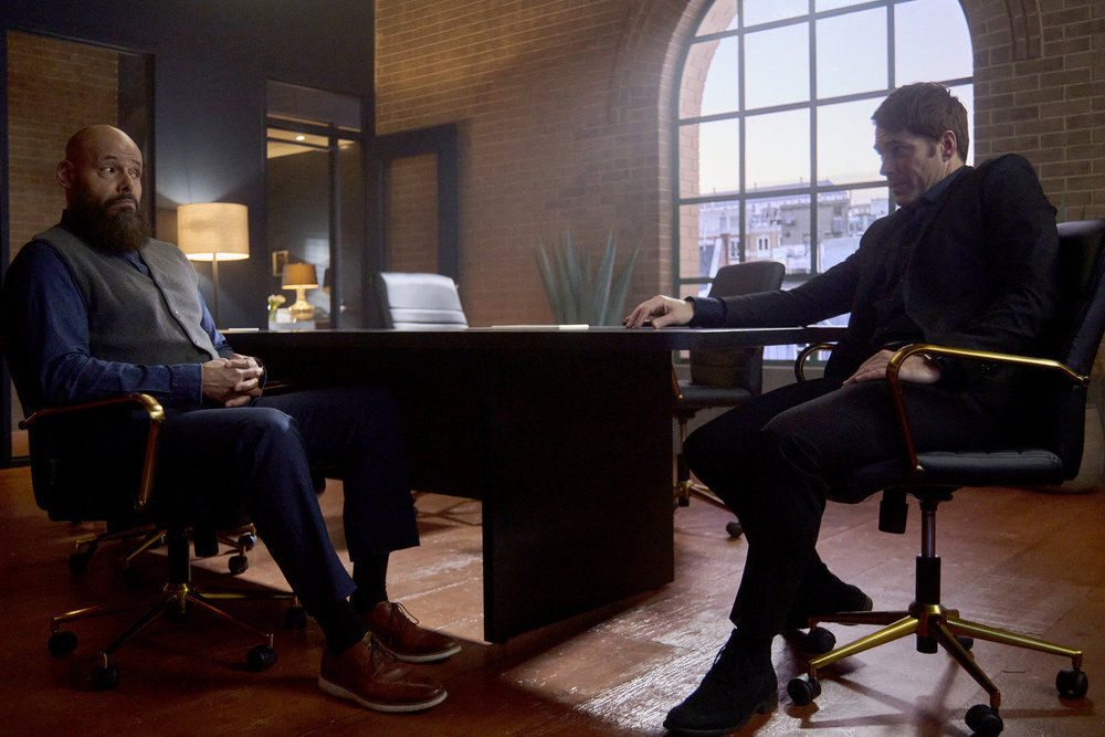 August and Luke sit at a table in a dimly lit office space in a real estate agency. They look at a screen that's out of frame on SurrealEstate Season 3 Episode 8, "Perchance to Dream."