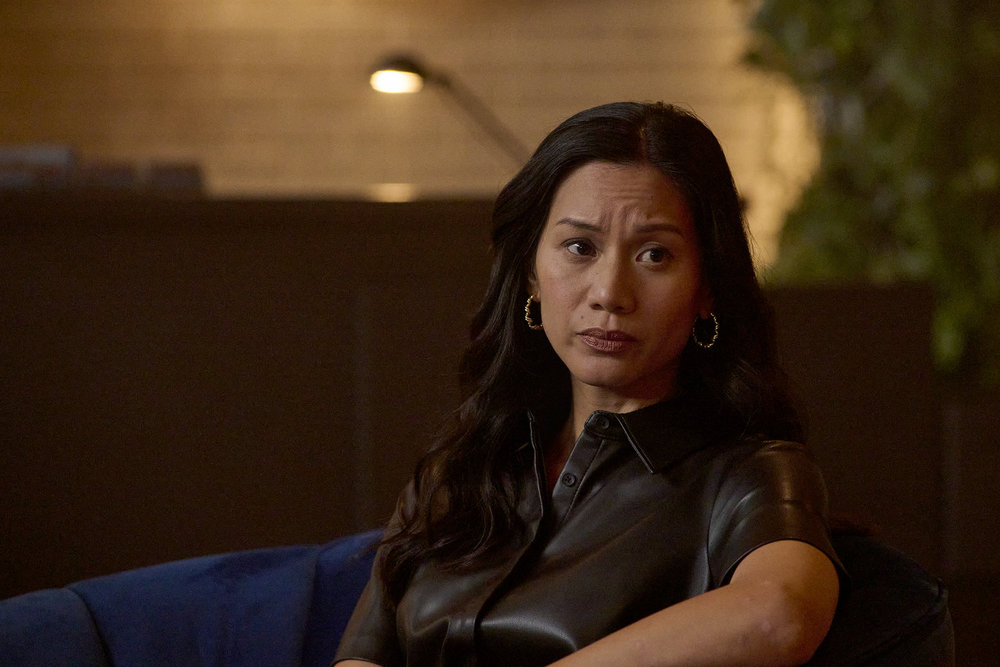 Lomax sits in a chair in a dimly lit office space in a real estate agency. She wears a black satin button-down short-sleeved top. She looks concerned on SurrealEstate Season 3 Episode 8, "Perchance to Dream."