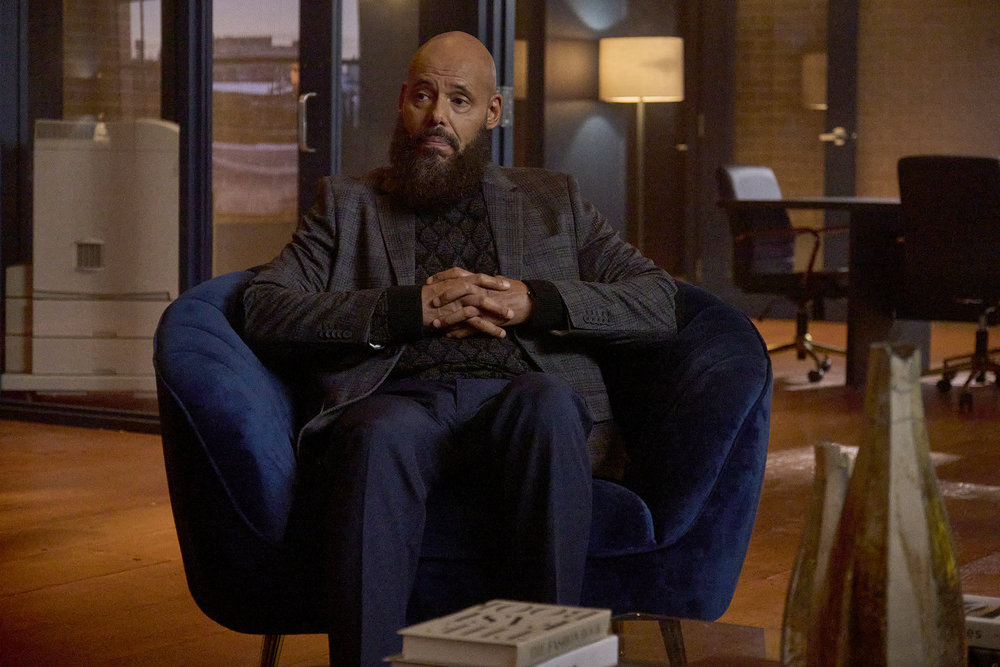 August, a bald Black man with a beard, sits in a navy blue sofa chair in a dimly lit office space in a real estate agency, his hands folded across his lap. He wears a gray suit jacket with matching pants and looks intently at something out of frame.