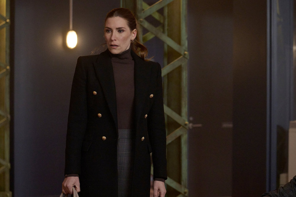 Susan, a white woman with auburn hair, stands in the entryway of a real estate agency. She wears a long black coat with a brown turtleneck long-sleeved shirt, her hair in a ponytail. She looks somber.