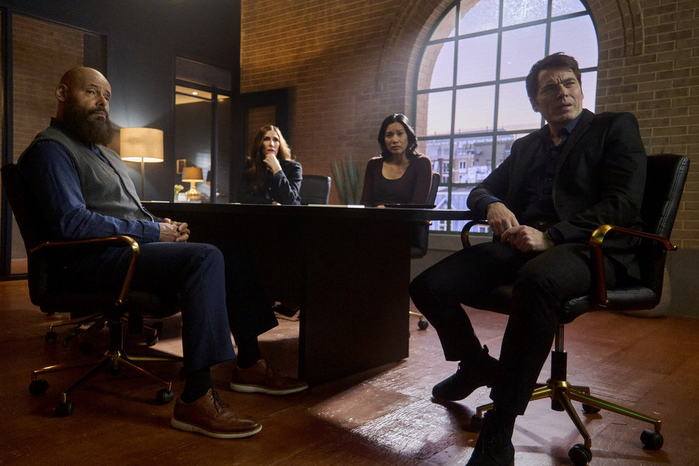 August, Susan, Lomax and Luke sit at a long table in their real estate agency while looking at a screen out of frame on SurrealEstate Season 3 Episode 8, "Perchance to Dream."