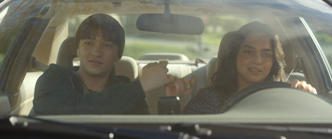 Charlie (Teo Briones) and Stefani (Kaitlyn Santa Juana) make a pinky promise while sitting in a car during the day.