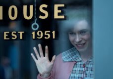 A white woman with brown hair waves and smiles creepily while standing before a store window in the TV series IT: Welcome to Derry.