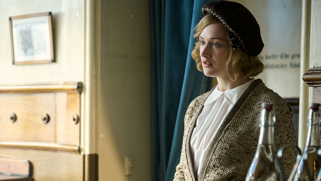 Unity stands facing to the left, dressed in a tweed jacket over a white blouse. She wears a tam hat. Behind her, blue drapes are pulled to the side, revealing a room with German words written on the wall in Outrageous Season 1 Episode 3.
