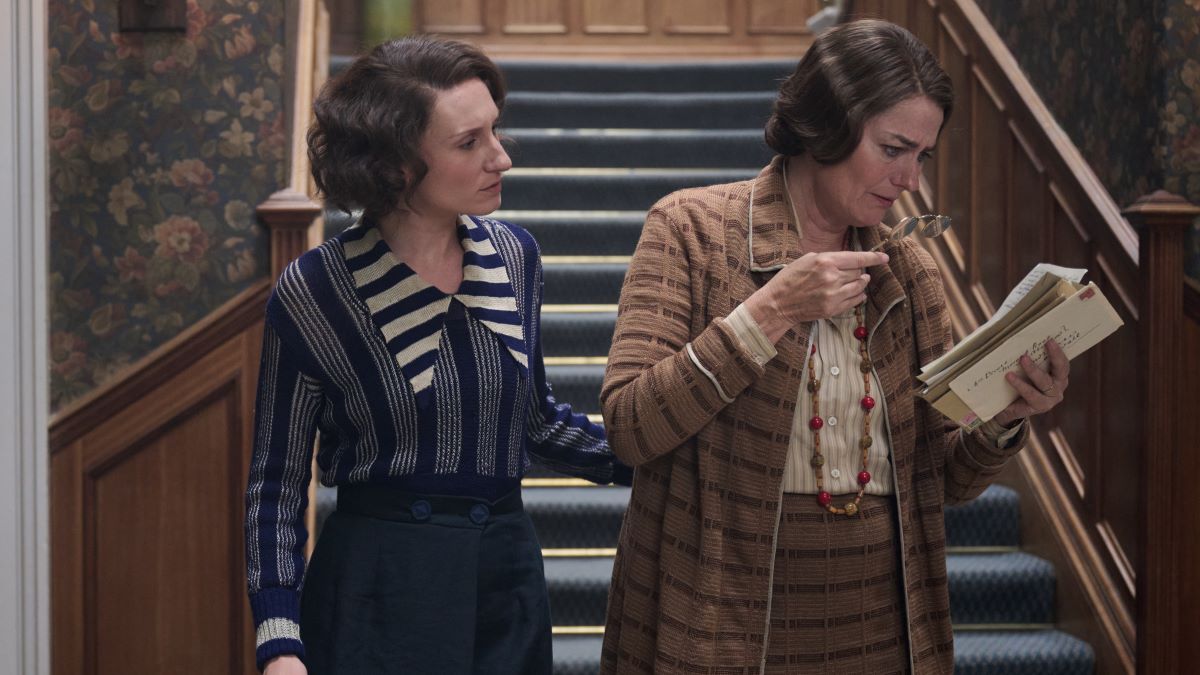 Nancy peers over Muv's shoulder as Muv reads a letter. Nancy wears a blue striped outfit, while Muv's dressed in a brown sweater and skirt coordinate set. Outrageous Episode 4