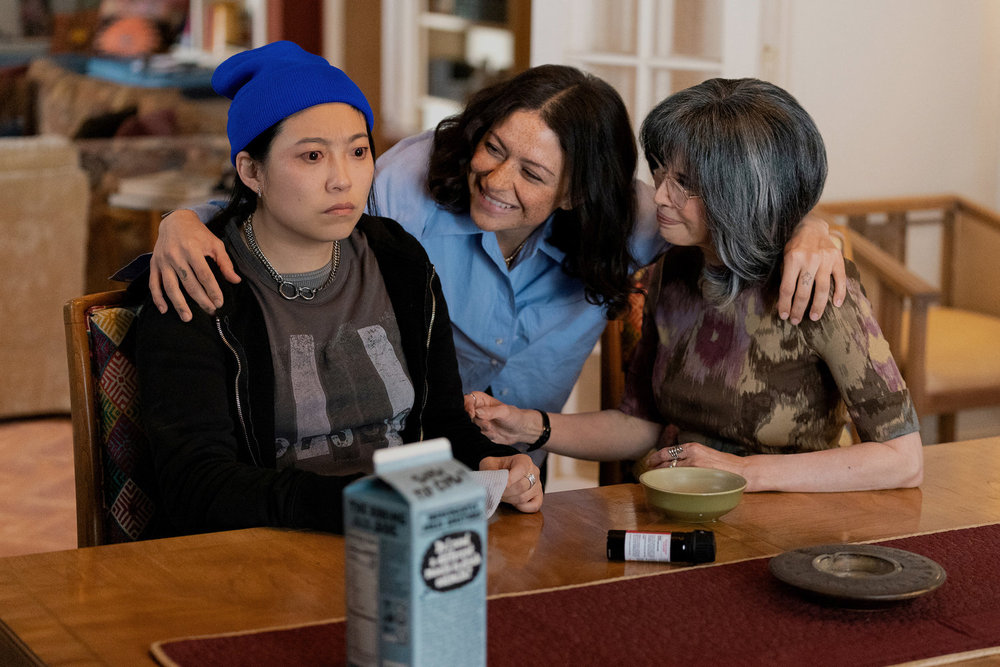 Maddy sits at a table in shock while Kate puts her arms around her. Maddy's grandmother, Anne, is on the other side of the table. All three are in a cozy penthouse in NYC on Poker Face Season 2 Episode 9, "A New Lease on Death."