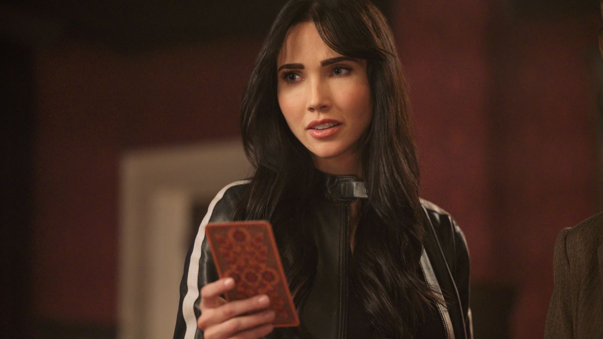Charlie holds a card up in her right hand. She's wearing a black leather jacket with a white stripe running down the sleeves. The Librarians: The Next Chapter Episode 106