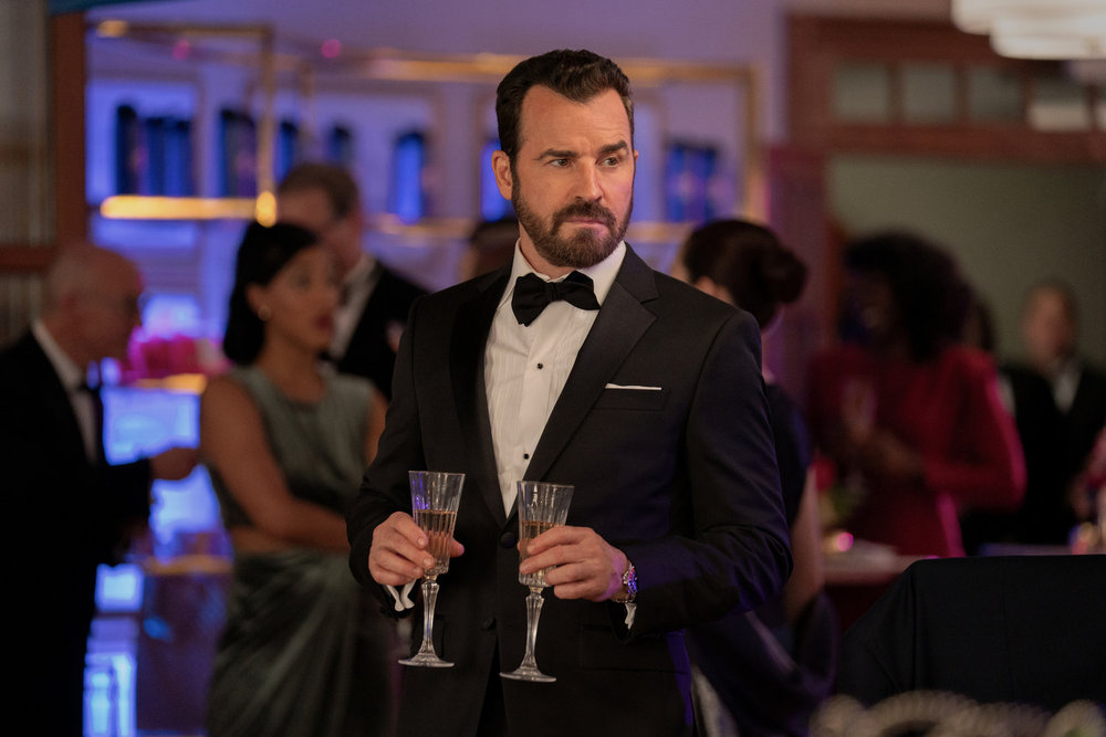 Iguana, a white man with brown hair and a beard, wears a suit while holding two champagne flutes and standing in the middle of a party on Poker Face Season 2 Episode 11, "Day of the Iguana."