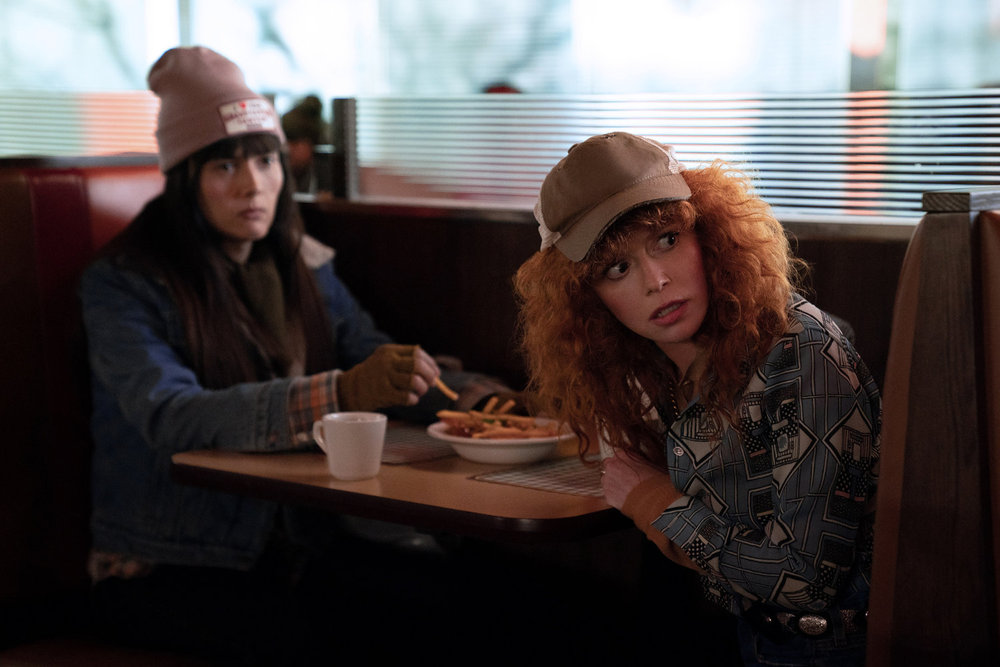 Alex and Charlie sit in a booth at a diner while looking at something out of frame on Poker Face Season 2 Episode 12, "The End of the Road."