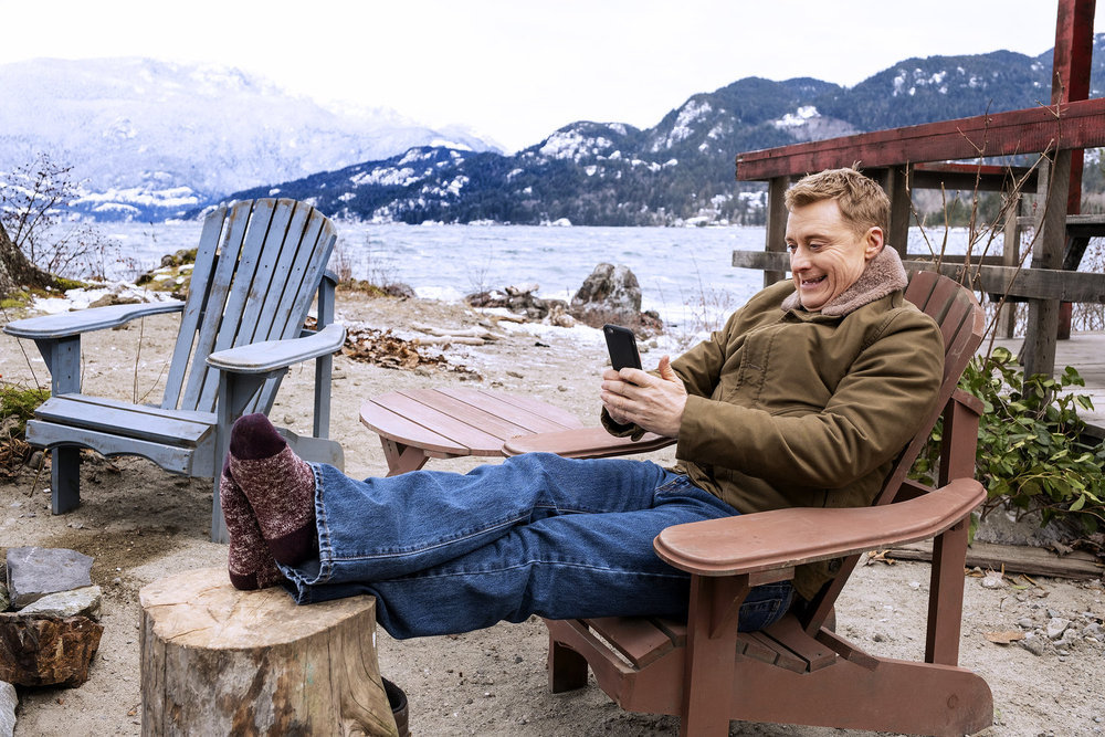Harry puts his feet up while sitting outside his cabin. He holds his phone to take a picture on Resident Alien Season 4 Episode 5, "The Human Condition."