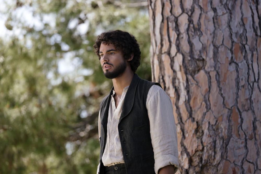 Guy stands outside in a park during the day, his back against a tree, while looking pensive on The Buccaneers Season 2 Episode 4, "Ice Cream."