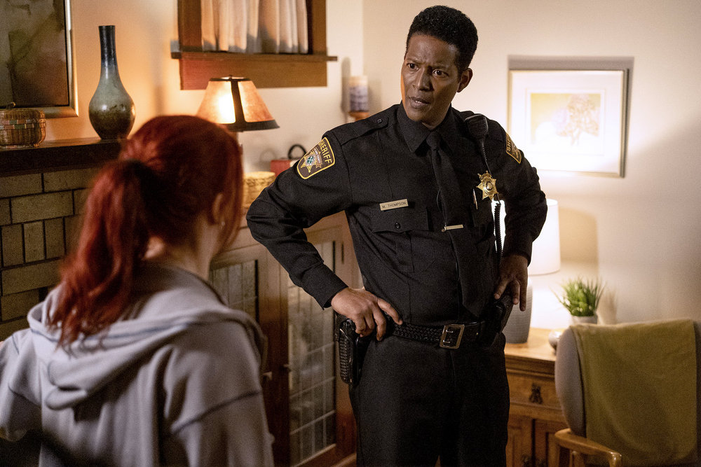 Sheriff Mike Thompson, a Black man in a sheriff's uniform, stands in a dimly lit living room of a small house. He has his hands on his hips while chatting with a white woman with bright red hair.