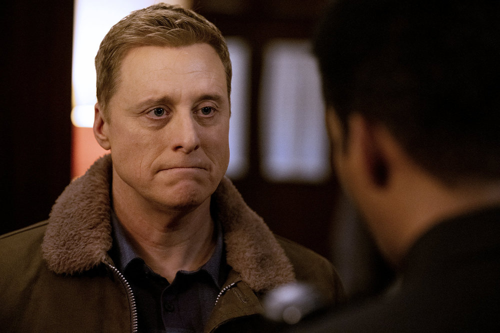 Harry, a middle-aged white man with short blonde hair, looks sad as he talks to Mike, a Black man, who's a sheriff. They stand in a dimly lit living room.